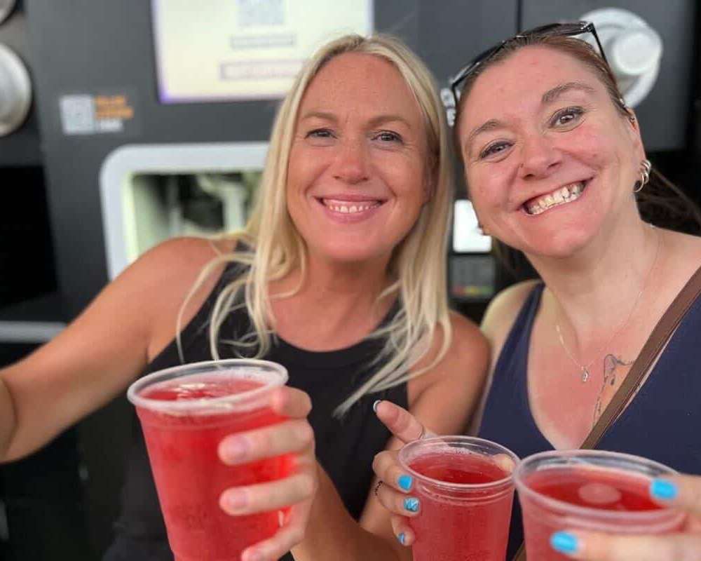 Two women smile for the camera while showing off the 4 pints of pink cider they have just received from the EBar units in the background