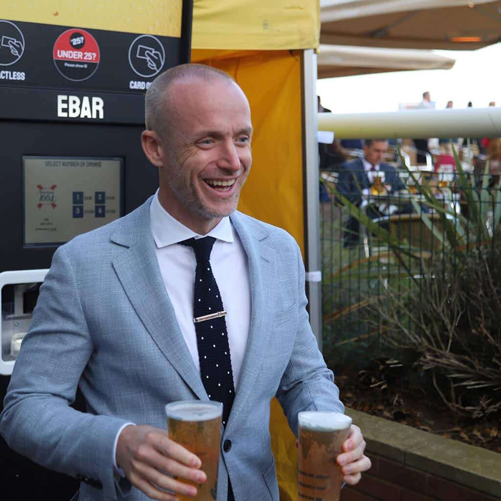 A smiling man in a suit walks away from an EBar automated beer kiosk , holding two pints of lager