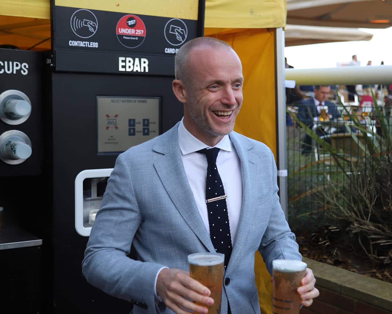 A smiling man in a suit walks away from an EBar unit, holding two pints of lager
