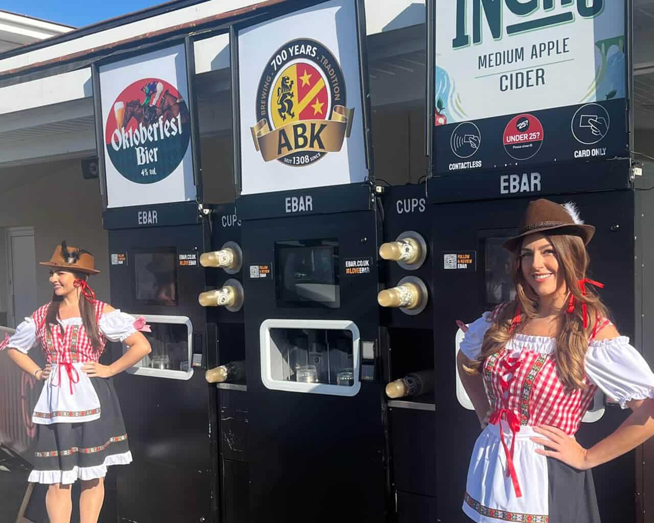 Two women in traditional Oktoberfest outfits stand smiling in front of 3 mobile EBar units