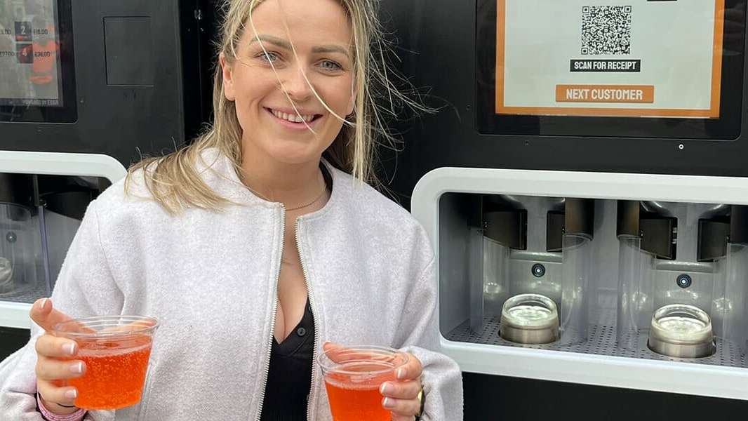 A smiling woman holds up two cocktails she just purchased from the EBar unit behind her