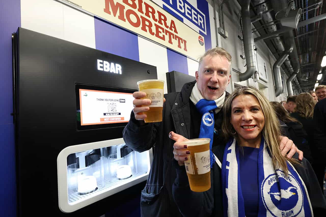 Two football fans smile to the camera after using the Beerwall behind them at the Amex Stadium, Brighton and Hove Albion FC